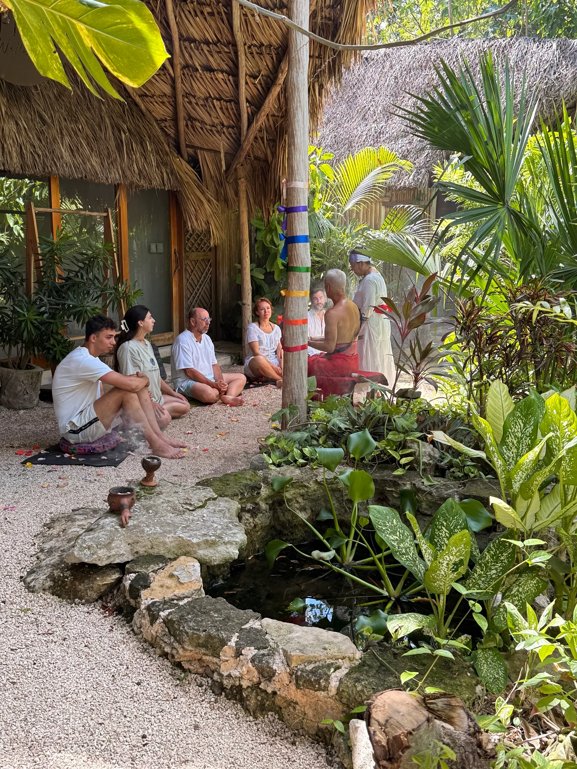 Un círculo de personas sentadas bajo la palapa, junto al poste de los listones